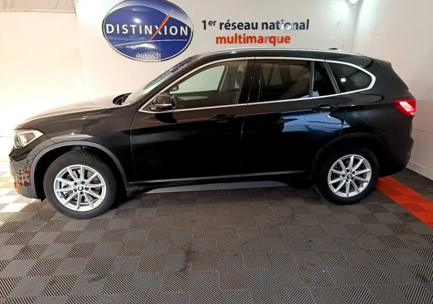 BMW X1 sDrive20i noir vue de profil côté gauche, stationné en intérieur avec logo Distinxion en arrière-plan.