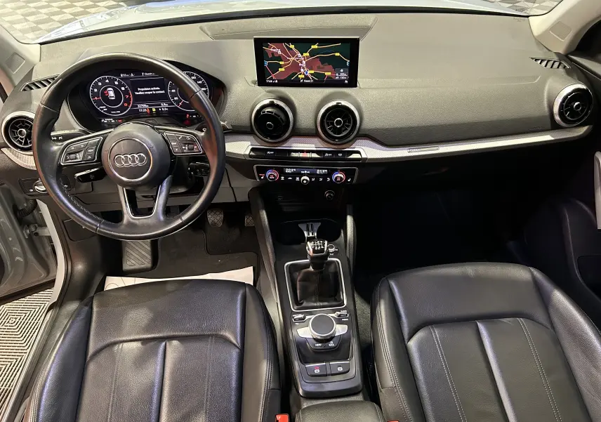 Vue intérieure avant de l'Audi Q2 gris, avec tableau de bord digital, écran GPS central et sièges cuir noirs.