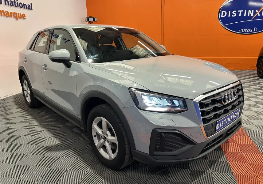 Audi Q2 gris en 3/4 avant droit, phares allumés et calandre hexagonale distinctive, dans un showroom intérieur.