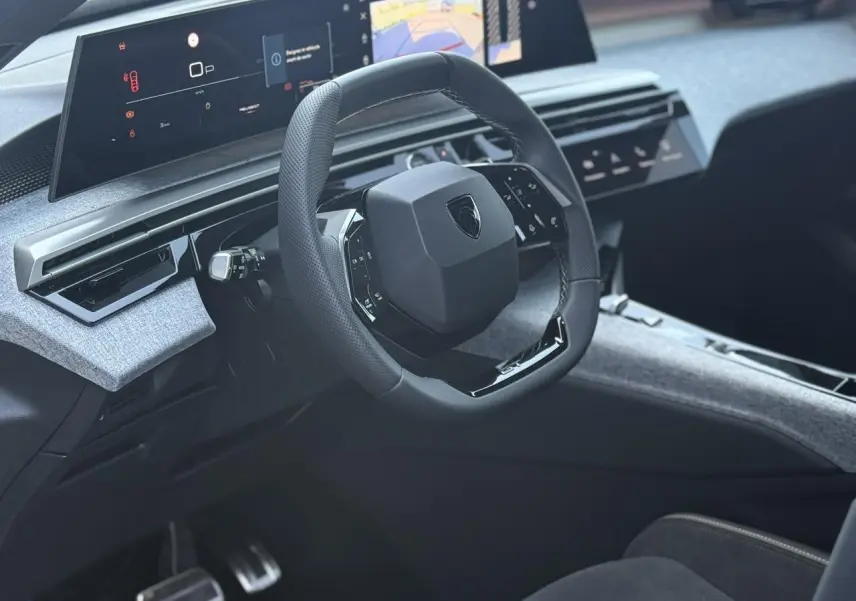 Vue intérieure du poste de conduite du Peugeot 5008 Hybrid 2026, volant compact noir laqué et tableau de bord numérique panoramique.