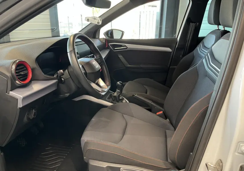 Intérieur côté conducteur d'une SEAT Arona blanc Nevada 2024, sièges textile noirs avec surpiqûres rouges et volant sport cuir.