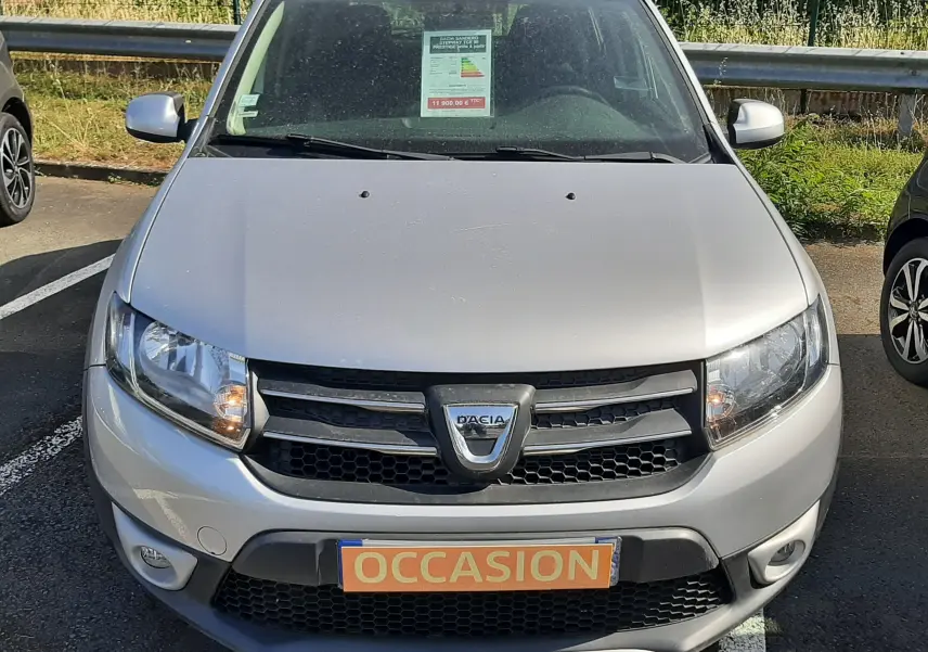 Vue frontale d'une Dacia Sandero Stepway gris platine avec plaque "OCCASION" sur un parking extérieur.