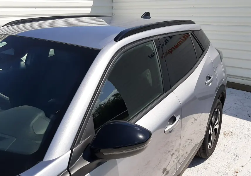 Vue de côté droit avant d’un Peugeot 2008 gris Artense avec barres de toit noires et vitres teintées.