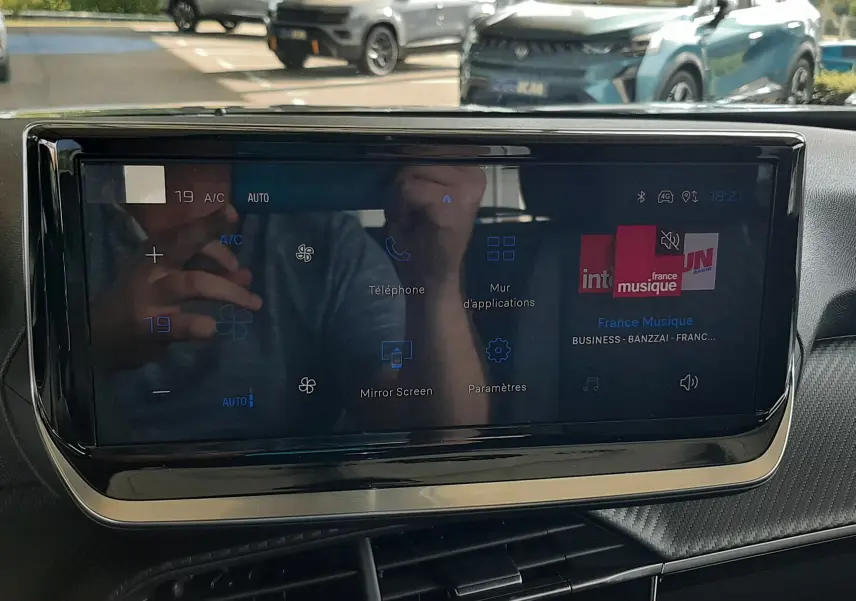 Vue intérieure centrée sur l'écran tactile du tableau de bord du Peugeot 2008, avec reflet d'un véhicule gris Artense à l'extérieur.