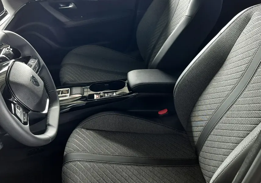 Vue intérieure côté conducteur du Peugeot 2008 1.2 Hybrid 145ch Allure 2025, sièges tissu noir avec surpiqûres et console centrale brillante.