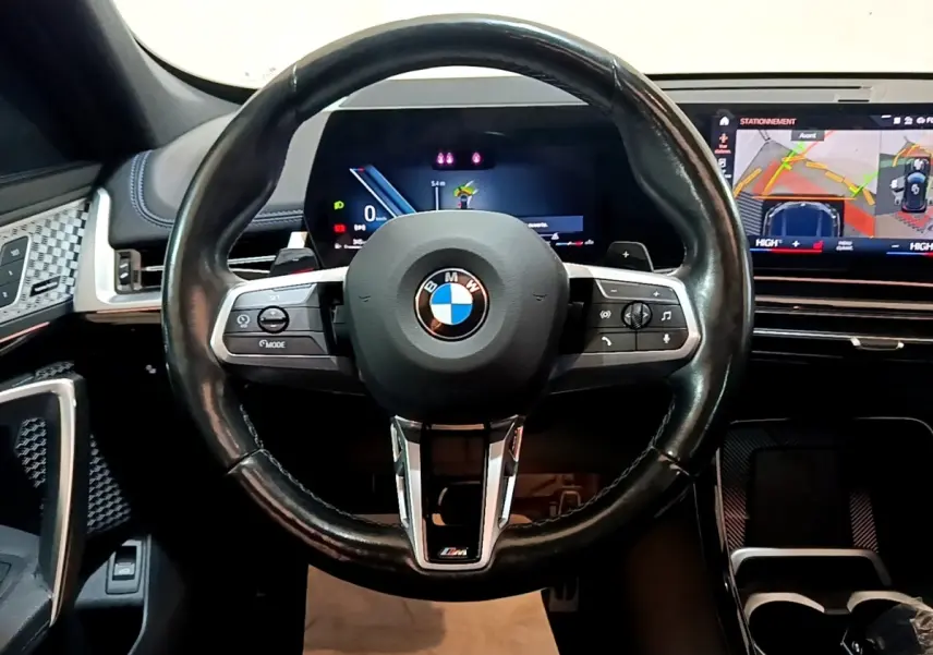 Vue intérieure centrée sur le volant cuir noir M Sport de la BMW X1 2023 avec tableau de bord digital et écran tactile.