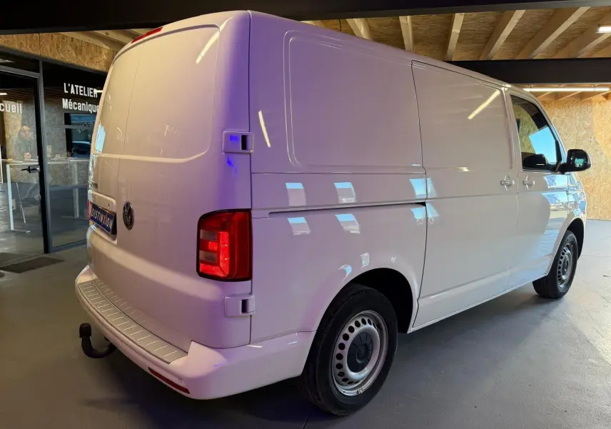 Vue 3/4 arrière droit d’un Volkswagen Transporter blanc 2019 en intérieur, avec feux arrière allumés et attelage visible.
