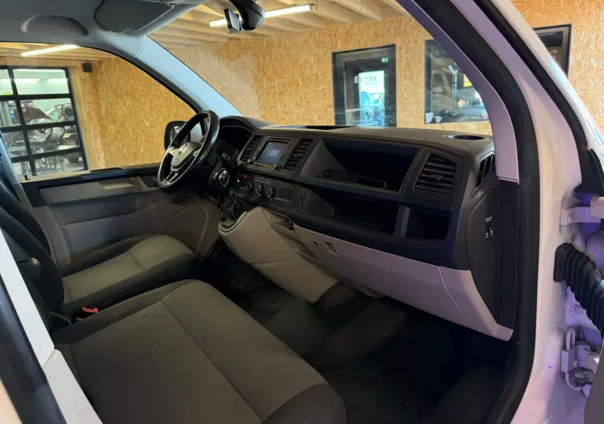 Vue intérieure côté conducteur du Volkswagen Transporter blanc 2019, tableau de bord et sièges en tissu noir visibles.