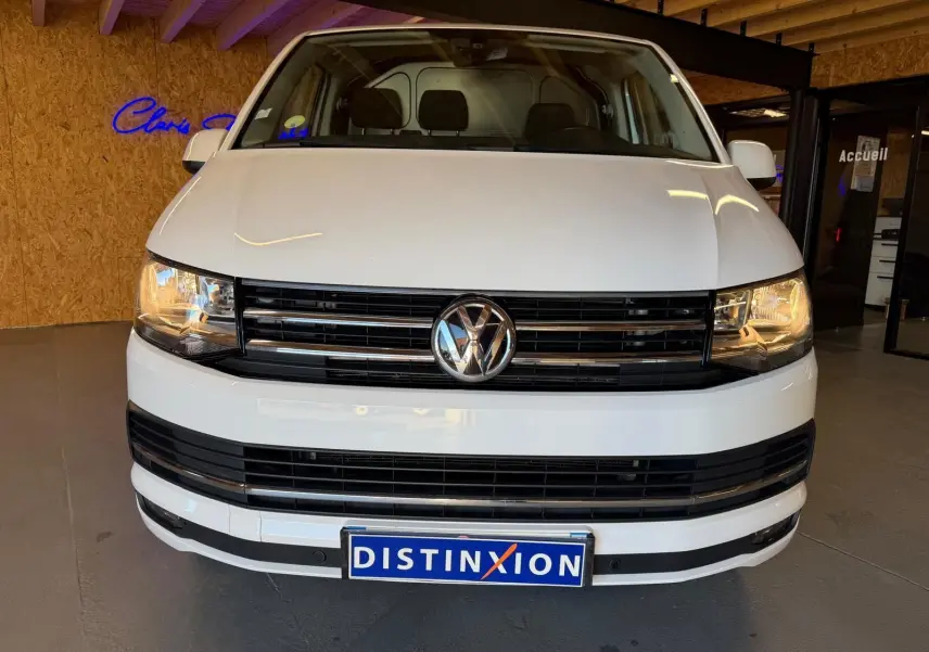 Vue frontale d’un Volkswagen Transporter blanc 2019 avec calandre noire et phares allumés en intérieur showroom.
