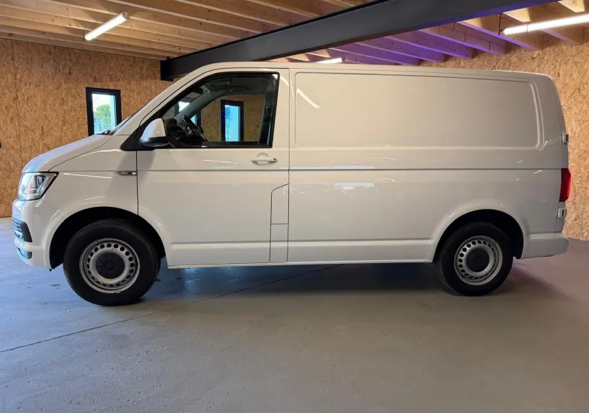 Vue de profil côté gauche d’un utilitaire Volkswagen Transporter blanc, stationné dans un garage avec murs en bois.