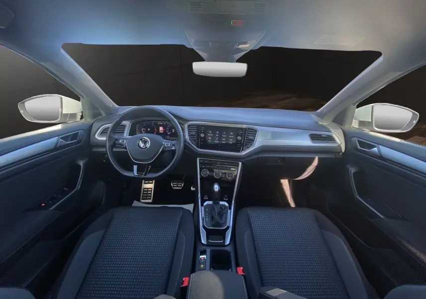 Vue intérieure avant du Volkswagen T-Roc 2021 avec tableau de bord noir, volant multifonction et toit ouvrant panoramique.