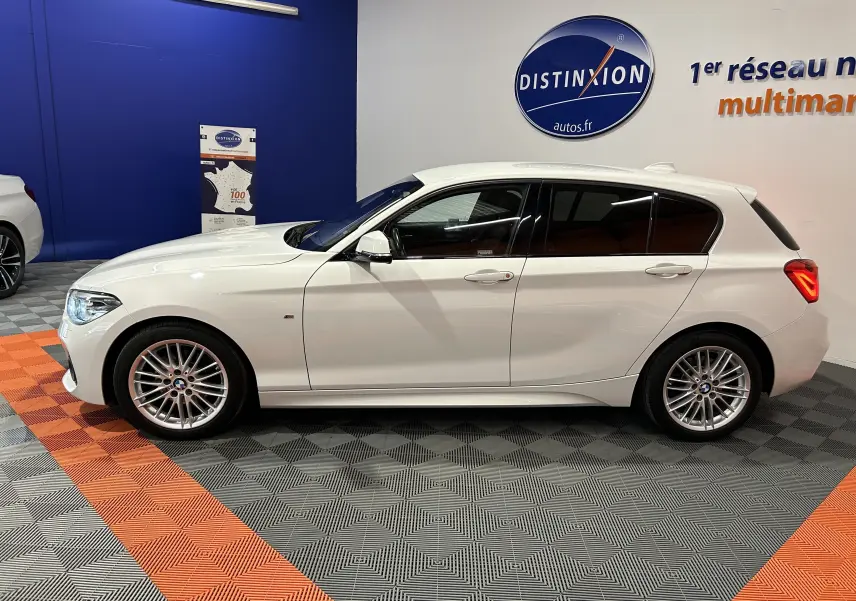 BMW Série 1 blanche vue de profil côté gauche, avec jantes alliage et vitres teintées dans un showroom.