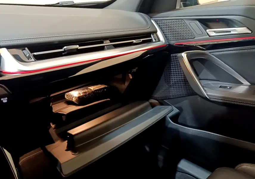 Vue intérieure côté passager du BMW X1 xDrive23i M Sport 2023, tableau de bord noir avec éclairage d'ambiance rouge et boîte à gants ouverte.