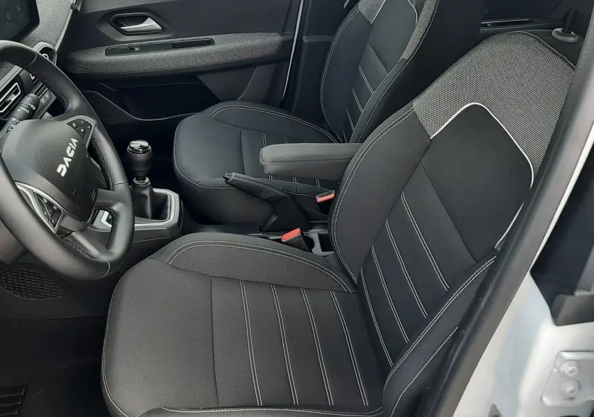 Vue intérieure côté conducteur de la Dacia Sandero 2024 blanc glacier, sièges tissu noir avec surpiqûres grises et volant multifonction.