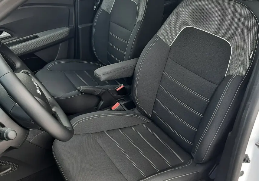 Vue intérieure côté conducteur de la Dacia Sandero 2024, sièges tissu noir avec surpiqûres blanches et accoudoir central.