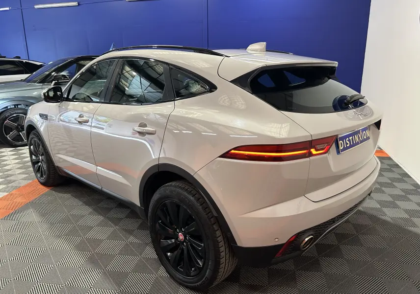 Jaguar E-Pace beige vue 3/4 arrière droit, feux arrière allumés, jantes noires et toit panoramique visible.
