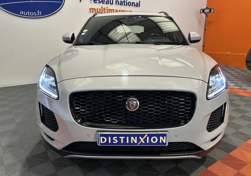 Vue frontale d'un Jaguar E-PACE beige avec calandre noire et phares LED allumés dans un showroom.
