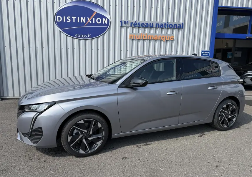 Peugeot 308 gris Artense vue de profil côté gauche, avec jantes noires et vitres teintées, devant un garage multimarque.