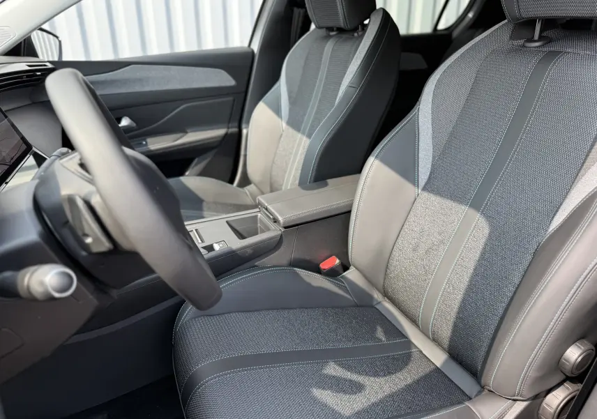 Intérieur de la Peugeot 308 2025, vue côté conducteur, sièges en tissu gris avec surpiqûres bleues et cuir noir.