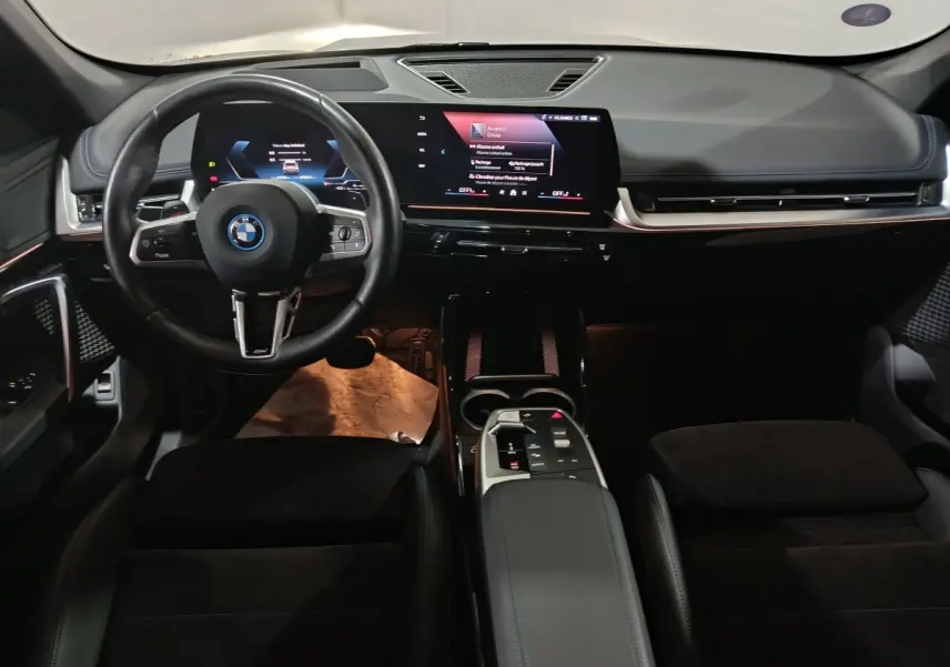 Vue intérieure avant du BMW X1 xDrive25e M Sport 2024, tableau de bord digital et volant cuir noir avec logo BMW.