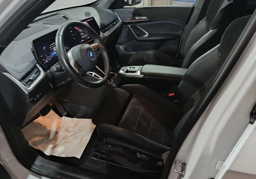 Vue intérieure côté conducteur du BMW X1 xDrive25e blanc 2024, avec tableau de bord digital et sièges sport noirs.