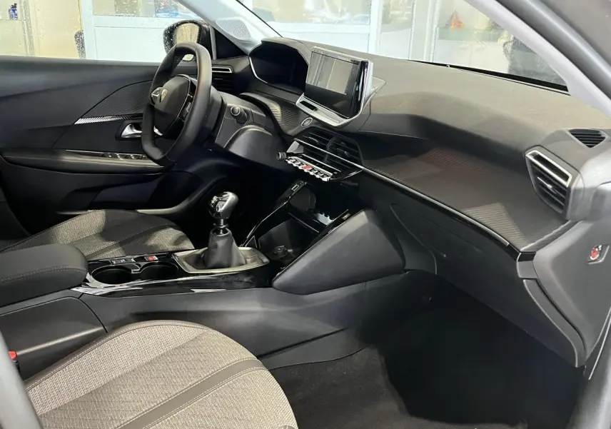 Vue intérieure côté conducteur de la Peugeot 208 2024, tableau de bord noir avec écran tactile et sellerie tissu gris clair.