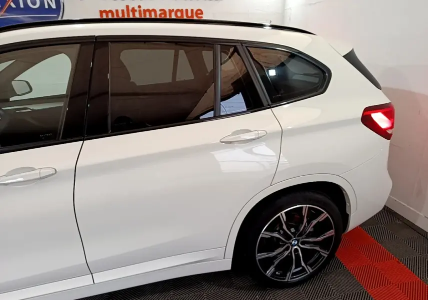 Vue de côté droit arrière d’un BMW X1 blanc avec jantes alliage noires et feux arrière allumés.