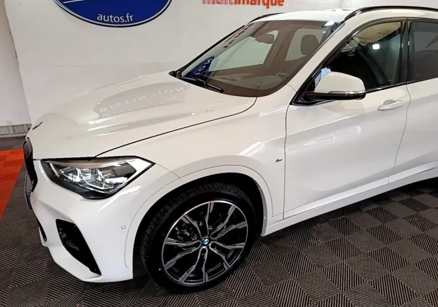 Vue 3/4 avant gauche d’un BMW X1 blanc sDrive18i M Sport avec jantes alliage noires et phares LED allumés.