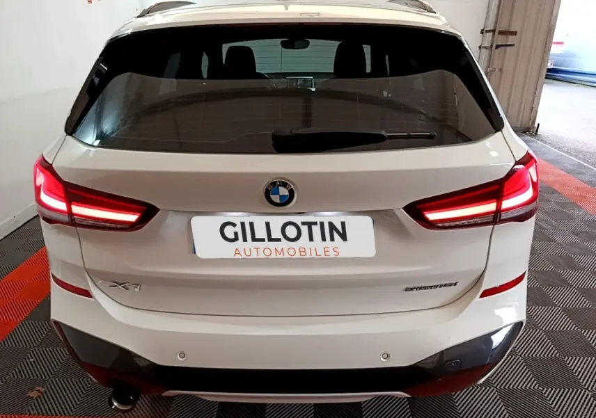 Vue arrière d’un BMW X1 blanc avec feux LED allumés, badge sDrive18i M Sport et double sortie d’échappement visible.
