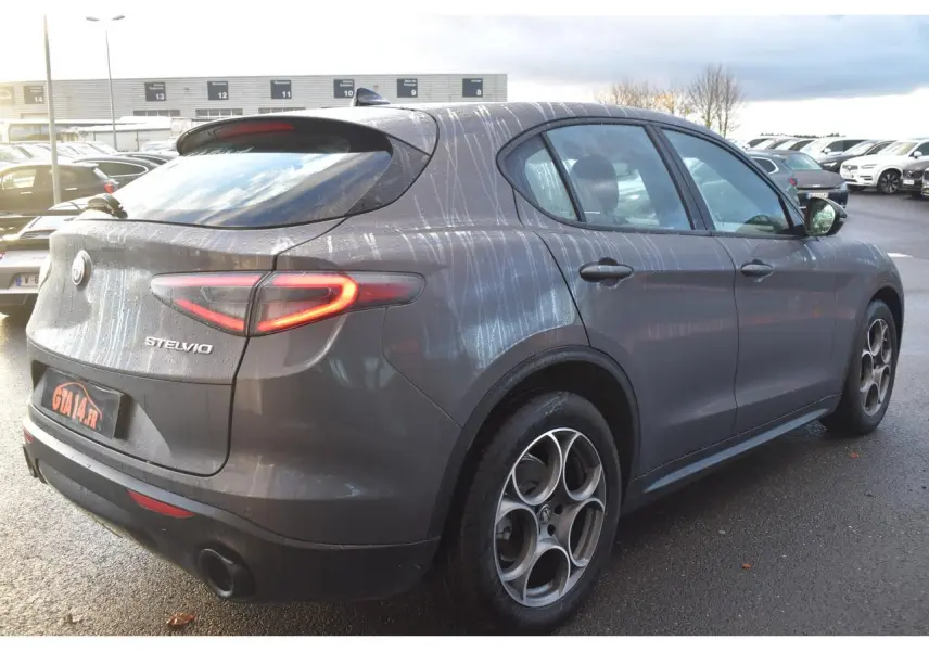 Vue 3/4 arrière droite d'un Alfa Romeo Stelvio 2024 gris Vesuvio avec jantes 19'' et feux arrière allumés.