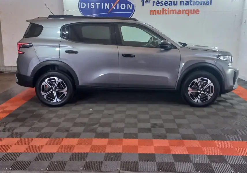 Vue latérale droite d'un Citroën C3 hybride gris foncé, stationné en intérieur sur sol damier noir et orange.