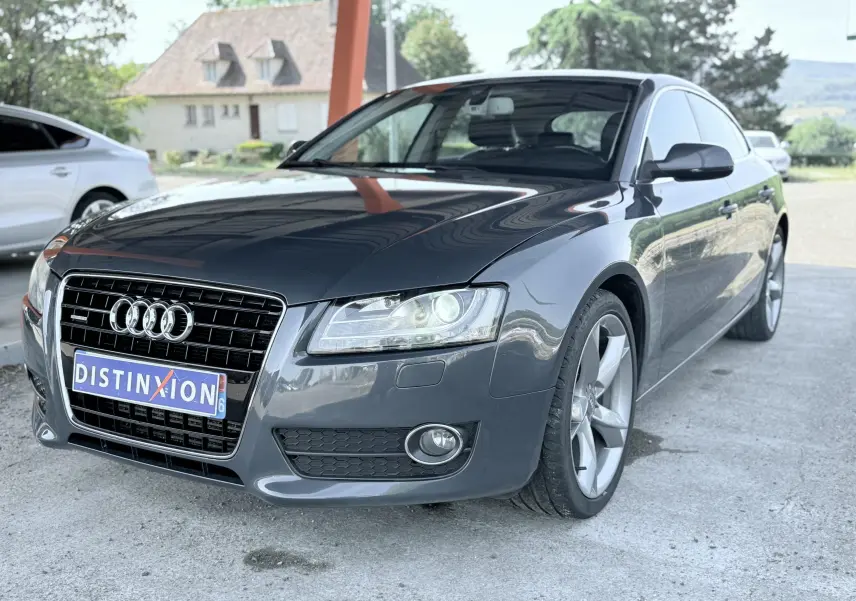 Audi A5 Sportback gris dauphin vue 3/4 avant droit, avec calandre noire et phares allumés sous abri.