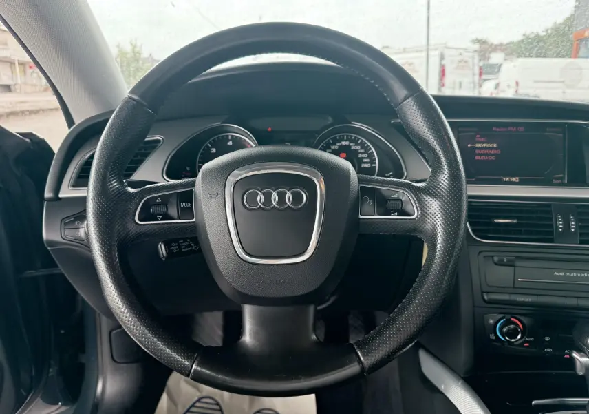 Vue intérieure centrée sur le volant en cuir noir perforé de l'Audi A5 Sportback avec tableau de bord et écran multimédia visibles.