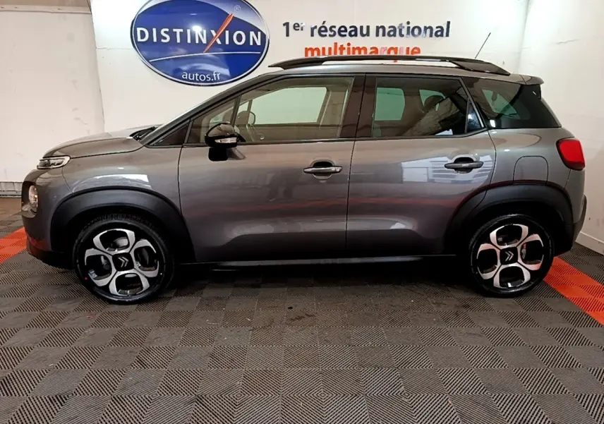 Vue de profil côté gauche d’un Citroën C3 Aircross gris foncé avec barres de toit noires en intérieur showroom.