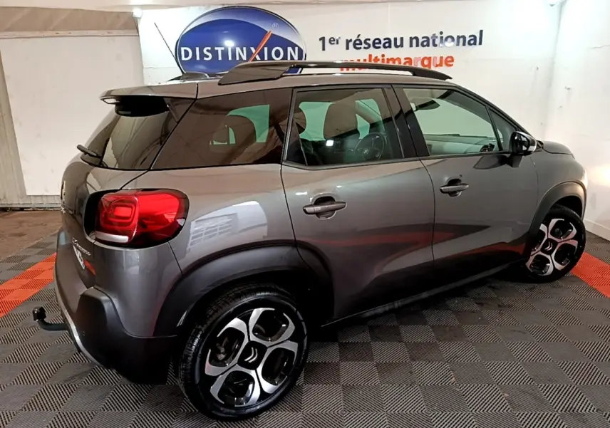 Vue 3/4 arrière droit d'un Citroën C3 Aircross gris foncé avec jantes bicolores et barres de toit noires.