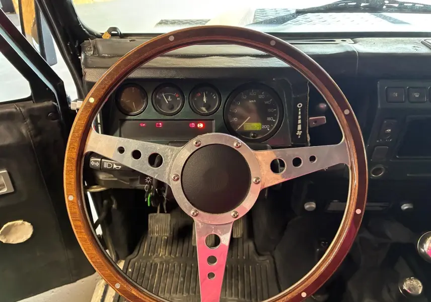 Vue intérieure centrée sur le volant bois du Land Rover Defender 110 vert de 2005 avec tableau de bord et pédales visibles.
