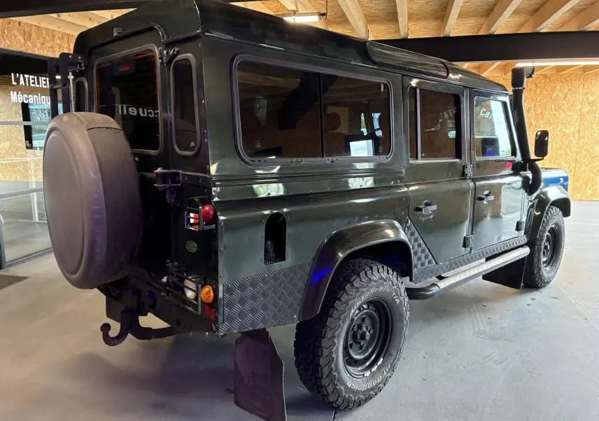Land Rover Defender 110 vert vu en 3/4 arrière droit, avec roue de secours et protections en métal sur les bas de caisse.