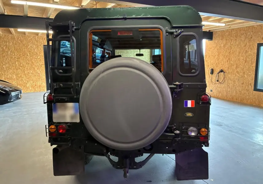 Vue arrière d’un Land Rover Defender 110 vert foncé de 2005, avec roue de secours et drapeau français sur la porte arrière.