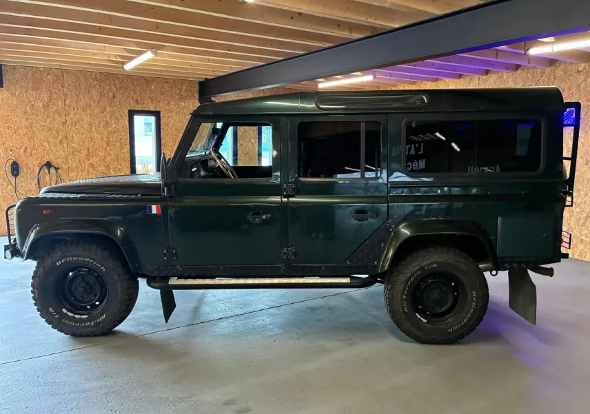 Vue de profil côté gauche d'un Land Rover Defender 110 vert foncé de 2005 avec pneus tout-terrain et drapeau français sur l'aile avant.