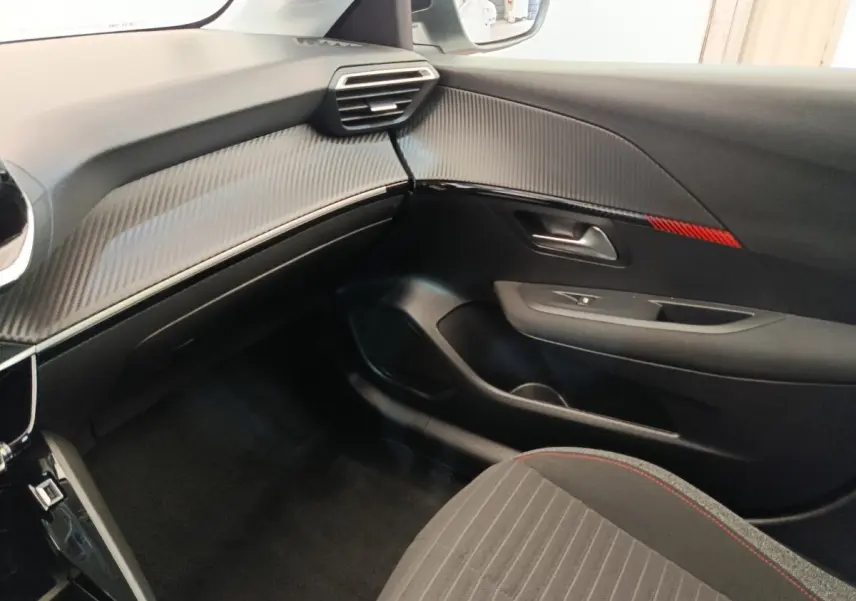 Vue intérieure côté passager de la Peugeot 208 blanche, tableau de bord avec insert carbone et bande rouge décorative.