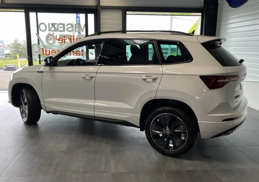 Profil droit du SUV SKODA KAROQ blanc 2025 avec jantes alliage noires et toit ouvrant visible en intérieur showroom.