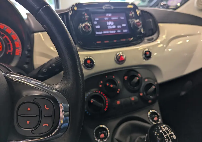 Vue intérieure du tableau de bord et volant de la Fiat 500 blanc Bossanova, avec commandes manuelles et écran central tactile.