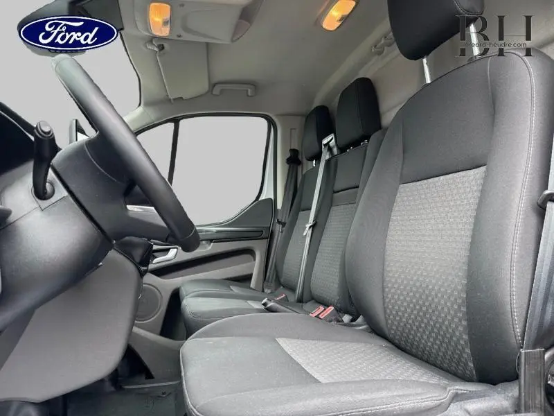 Vue intérieure côté conducteur du Ford Transit Custom Fourgon 2023 avec sièges tissu gris et volant cuir noir.