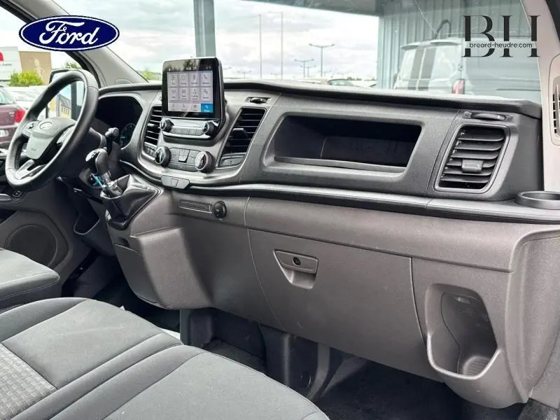 Intérieur du Ford Transit Custom blanc, vue côté passager montrant tableau de bord avec écran tactile et sièges tissu gris.