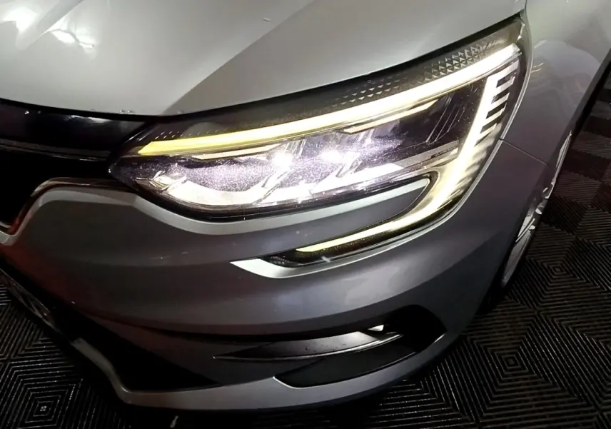 Gros plan sur le phare avant droit allumé de la Renault Mégane Business Blue dCi 115 EDC gris clair, vue en légère plongée.