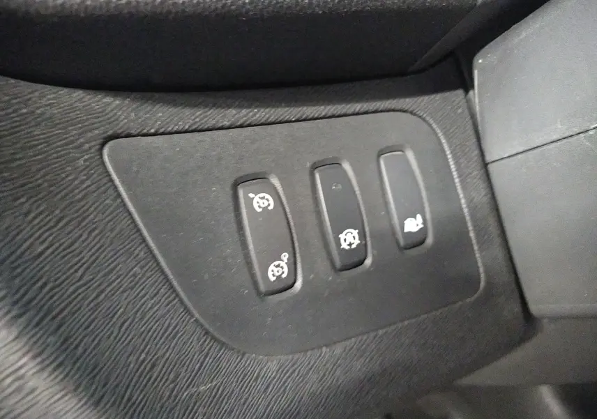 Gros plan sur les commandes noires du tableau de bord du Renault Kangoo blanc, avec symboles de régulateur et ouverture du coffre.