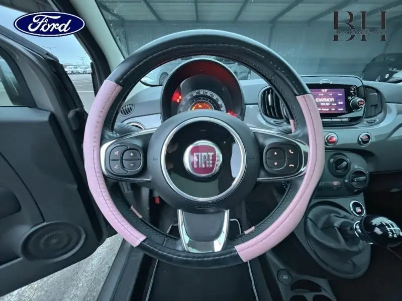 Vue intérieure centrée sur le volant cuir noir et rose de la Fiat 500 avec tableau de bord et écran tactile visibles.