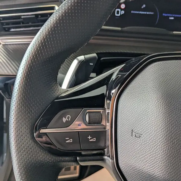 Gros plan sur le volant cuir noir perforé de la Peugeot 508 SW 2024, avec commandes audio intégrées et tableau de bord numérique en arrière-plan.