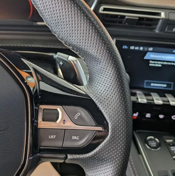 Gros plan sur le volant cuir perforé noir de la Peugeot 508 SW 2024 avec commandes audio et tableau de bord gris noir.