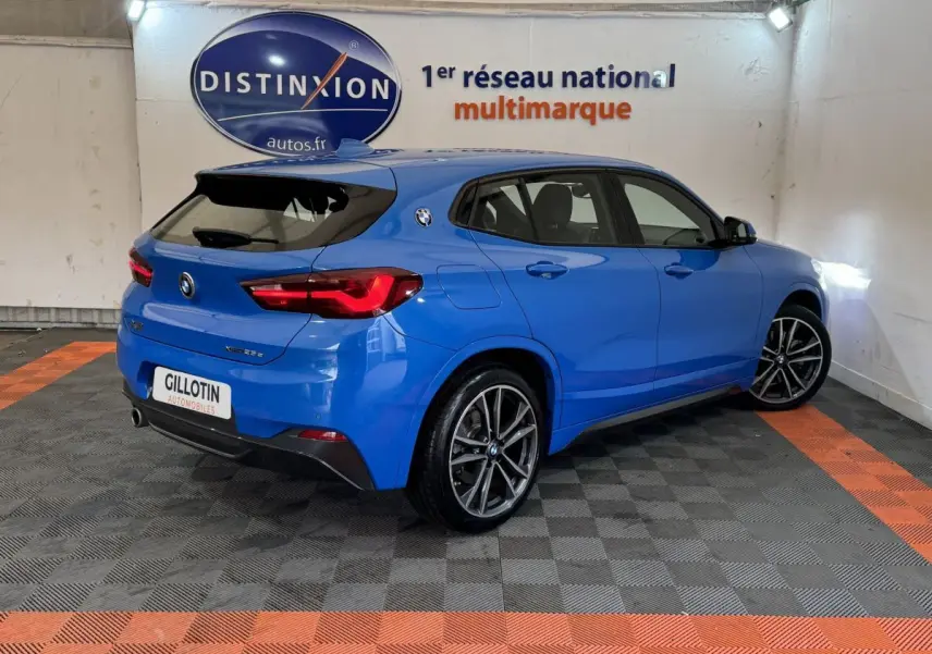 BMW X2 xDrive25e bleu en 3/4 arrière droit dans un showroom avec jantes sport et logo BMW sur l'aile arrière.